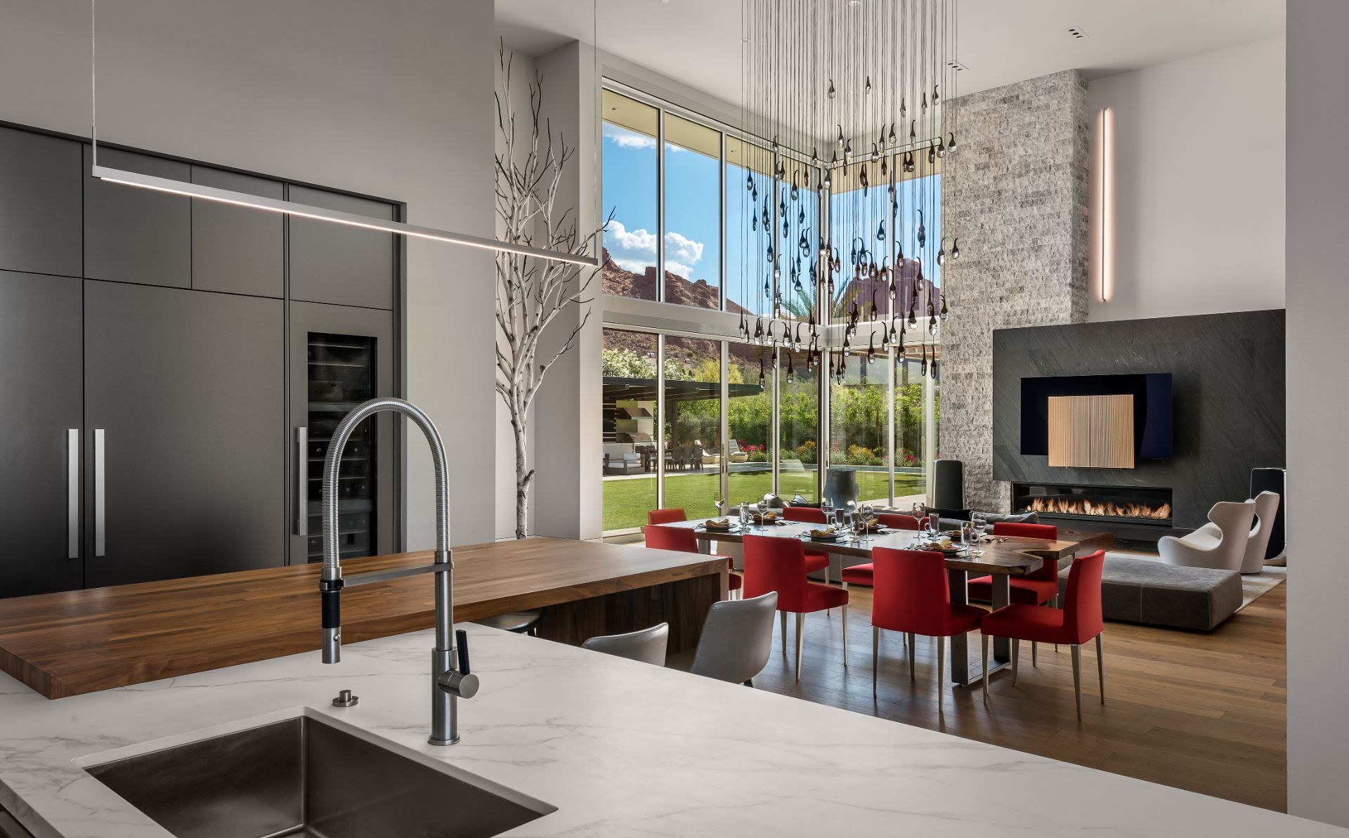 Open-plan kitchen and dining area of a Scottsdale project home, floor-to-ceiling glass framing Camelback Mountain, with a media wall above a linear fireplace